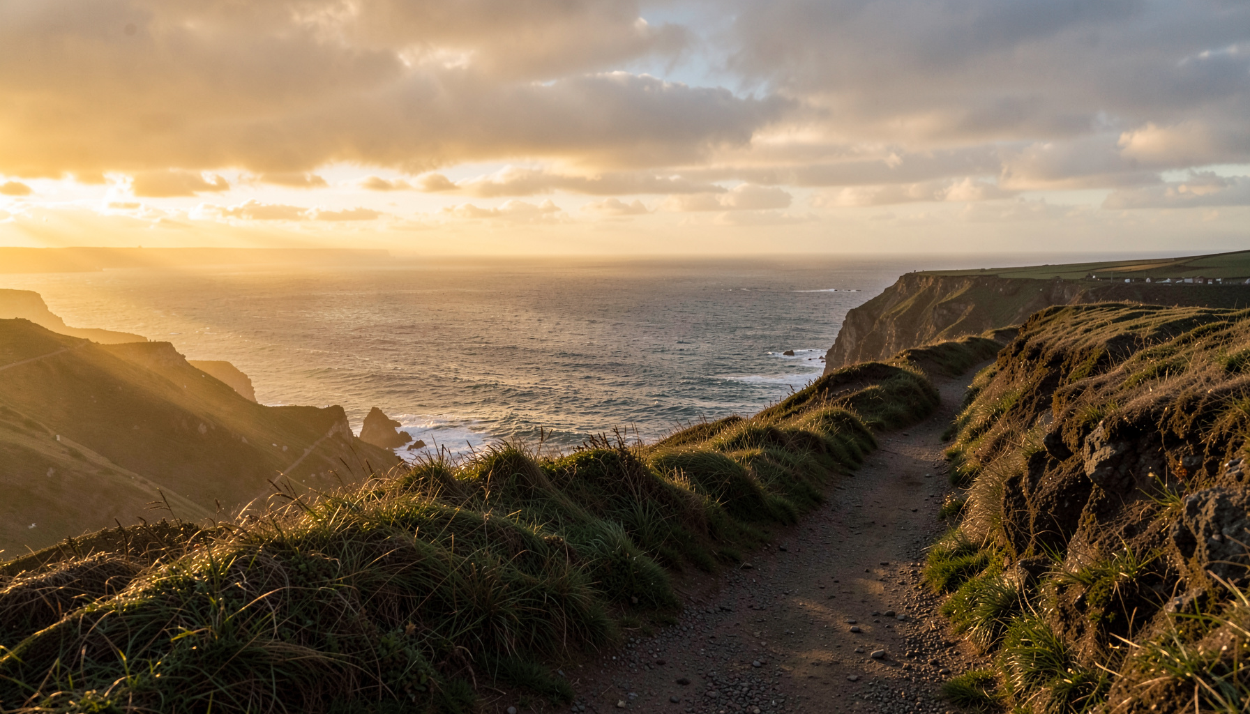 A windswept coastal cliff path overlooking the English Channel at dusk, with soft golden light breaking through clouds over the sea